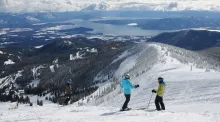Three skiers standing atop a snow-covered mountain slope overlooking pine forests and Lake Pend Oreille under a partly cloudy sky.