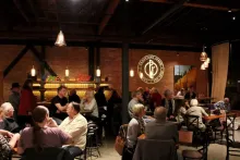 Guests gather at tables inside the Pend d'Oreille Winery tasting room in Sandpoint, beneath its illuminated winery logo on a brick wall.