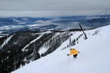 A skier in a yellow jacket descends a snowy Schweitzer slope beside a T-bar lift, with Lake Pend Oreille spread out below the mountain.