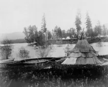 Historic photograph of a Kalispel tule-mat lodge and canoe on the shore of the Pend Oreille River