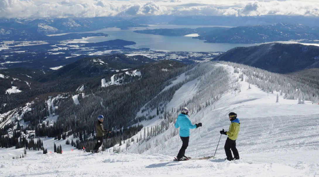 Three skiers standing atop a snow-covered mountain slope overlooking pine forests and Lake Pend Oreille under a partly cloudy sky.