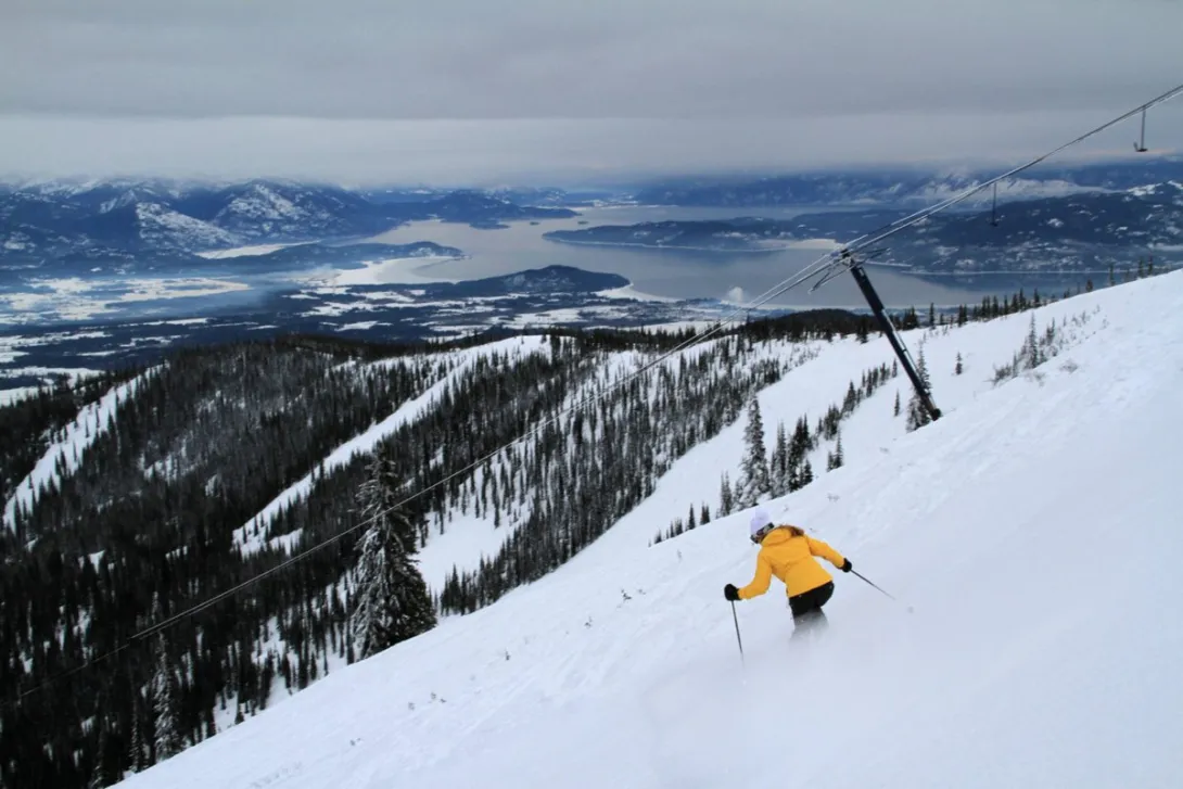 A skier in a yellow jacket descends a snowy Schweitzer slope beside a T-bar lift, with Lake Pend Oreille spread out below the mountain.