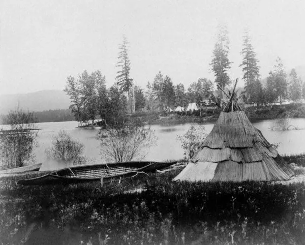 Historic photograph of a Kalispel tule-mat lodge and canoe on the shore of the Pend Oreille River